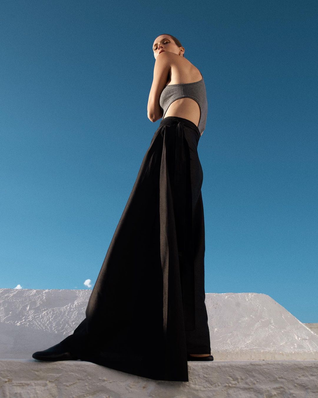 Fashion shot of a woman in a grey bodysuit and wide black pants standing on a white structure against a clear blue sky backdrop.