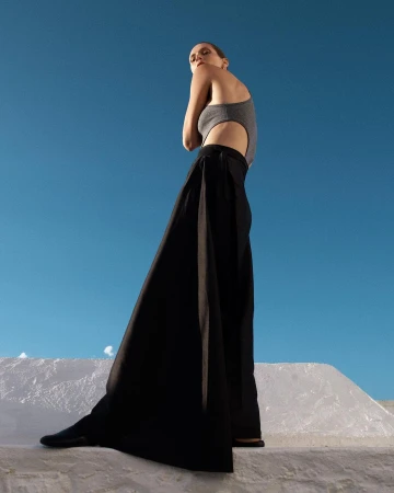 Fashion shot of a woman in a grey bodysuit and wide black pants standing on a white structure against a clear blue sky backdrop.