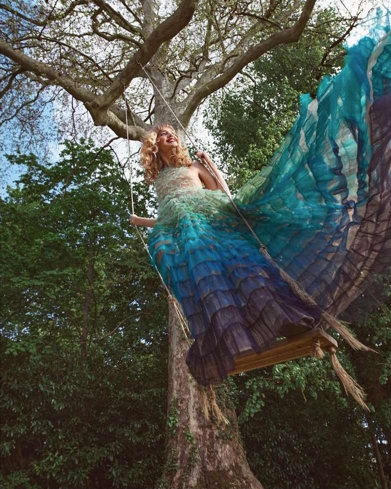 A woman in a long flowing blue dress is sitting on a swing that is hung from a large tree. The sky is visible behind the tree.