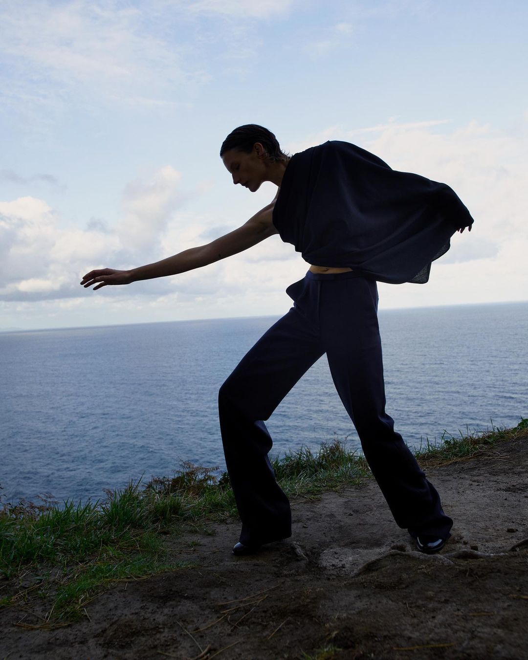 Fashion image of an adult female model in dark, loose clothing, posing on a grassy cliff with the ocean and sky in the background.