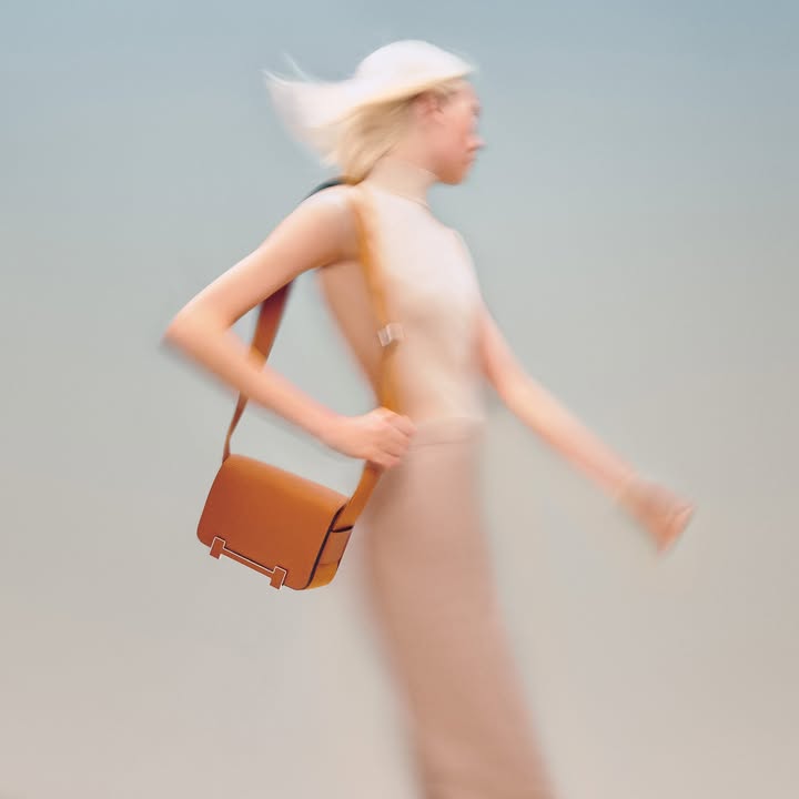 Fashion shot of a blonde woman with a tan leather purse slung over her shoulder, walking against a light grey background in motion blur.