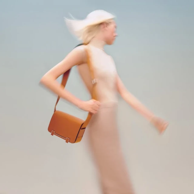 Fashion shot of a blonde woman with a tan leather purse slung over her shoulder, walking against a light grey background in motion blur.