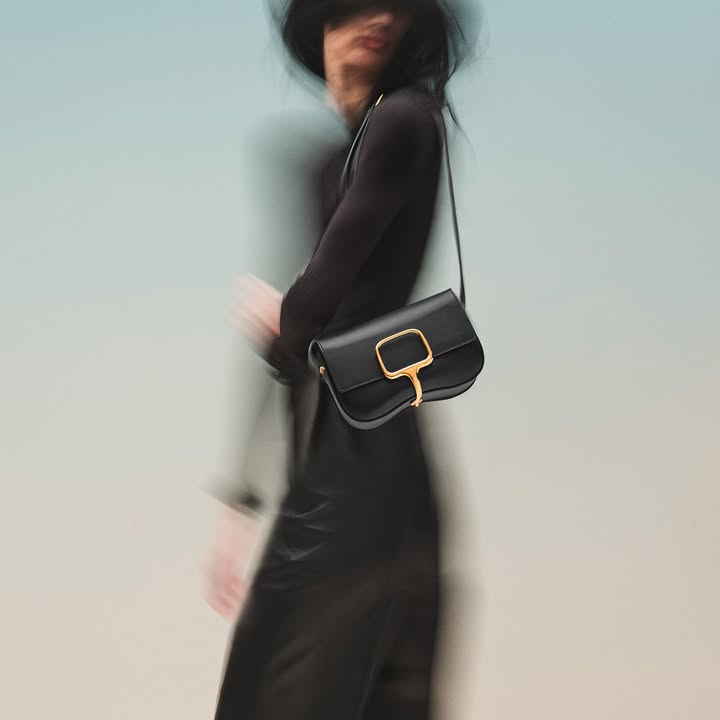Model wearing a black dress holding a black leather shoulder bag with a gold clasp against a blurred backdrop.