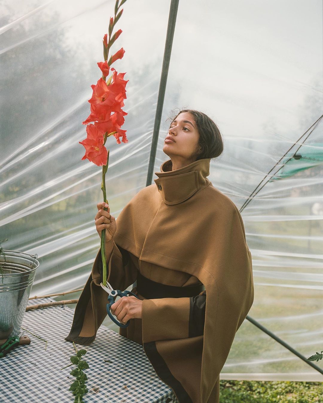 Young woman in a tan poncho trimming red gladiolus flowers inside a greenhouse, captured in a fashion editorial style photograph.