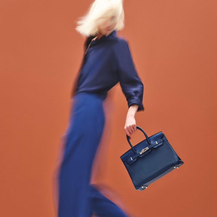 Blonde model in motion wearing blue shirt and pants holding a blue handbag against an orange background