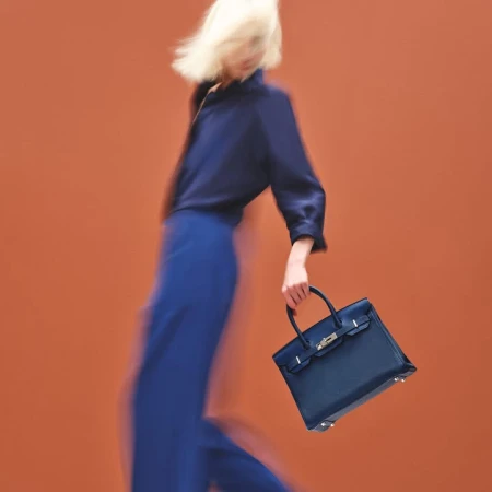 Blonde model in motion wearing blue shirt and pants holding a blue handbag against an orange background