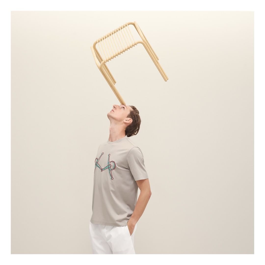 Man in a grey t-shirt with a bicycle print looking up at a light-wood chair balanced on his head against a neutral backdrop.