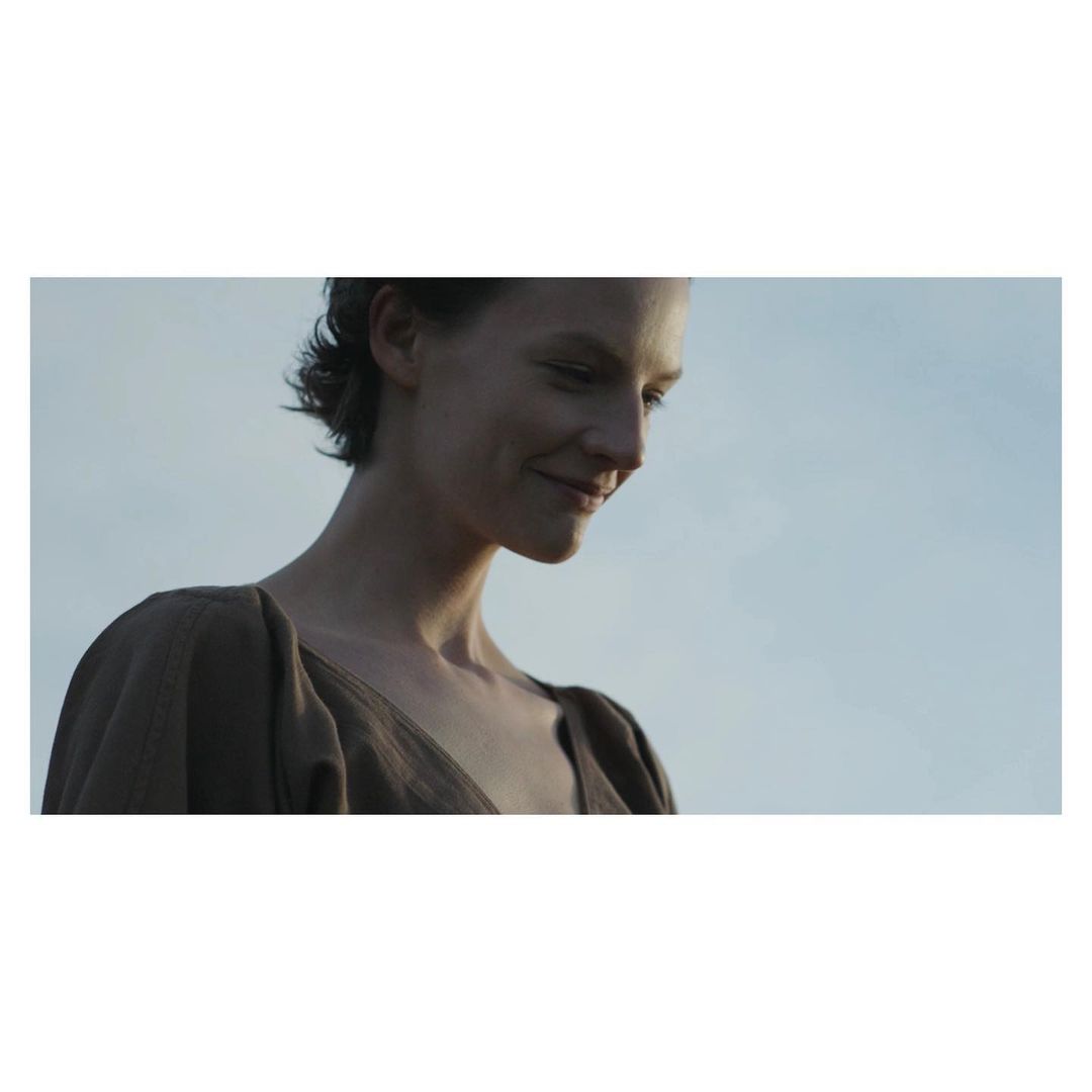 Portrait of a pensive adult female with short dark hair wearing a brown dress, captured in natural light against a soft blue sky.