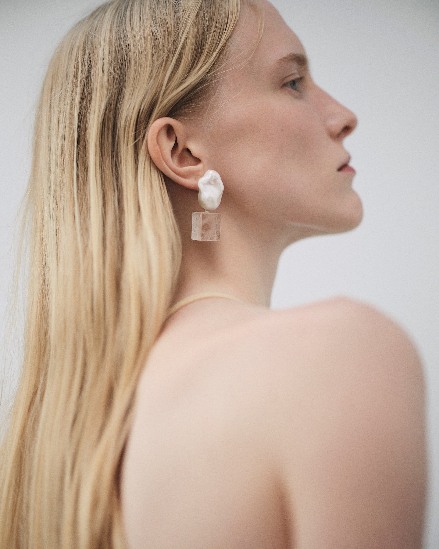 Portrait of a blonde adult female model with fair skin wearing distinctive geometric earrings in a neutral photo studio setting.