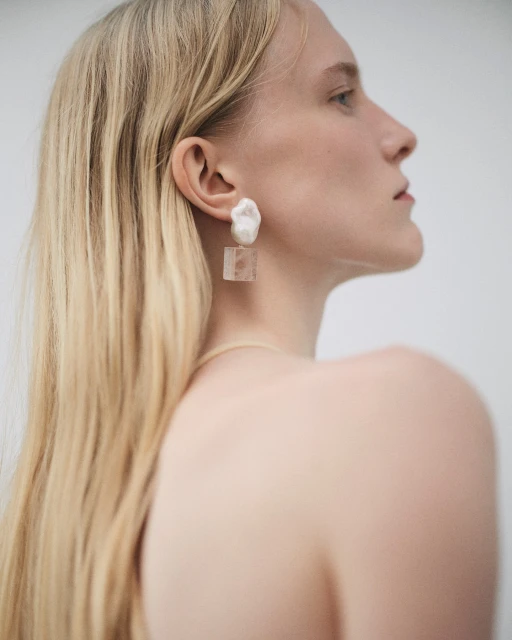 Portrait of a blonde adult female model with fair skin wearing distinctive geometric earrings in a neutral photo studio setting.