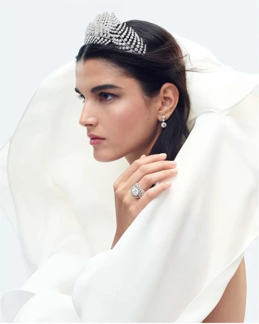 Woman with dark hair wears a diamond tiara, diamond earrings, and a large diamond ring, posed against a white draped fabric background.
