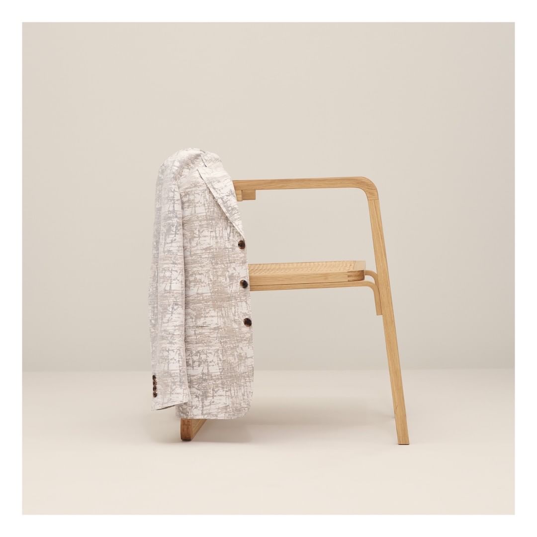 White blazer with a subtle pattern hangs on a light-wood chair against a cream-colored backdrop in a softly lit, staged photograph.