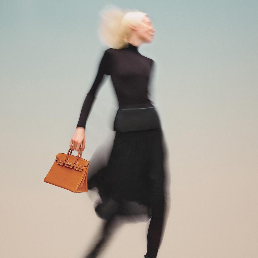 Fashion shot of a slender adult female with blonde hair wearing black, carrying an orange leather handbag, blurred motion effect.