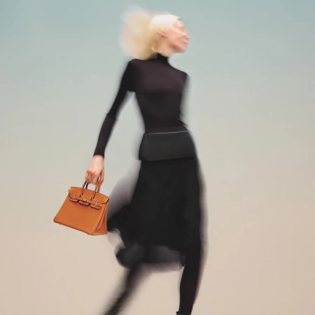 Fashion shot of a slender adult female with blonde hair wearing black, carrying an orange leather handbag, blurred motion effect.