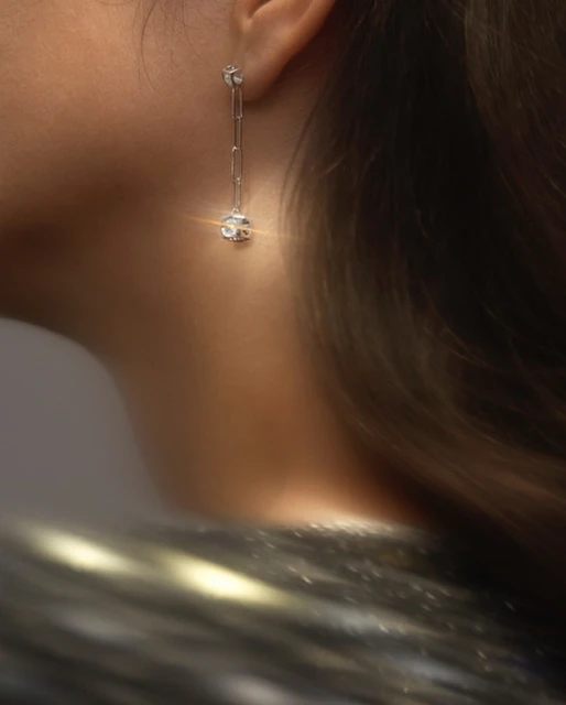 Elegant long silver chain earrings worn by a woman, close-up capturing ear and neck details with blurred textured clothing.
