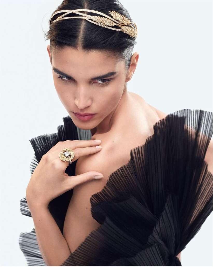Model wearing gold leaf headpiece and statement ring with black pleated dress against white background