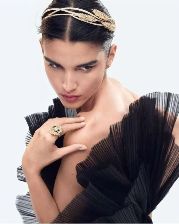 Model wearing gold leaf headpiece and statement ring with black pleated dress against white background