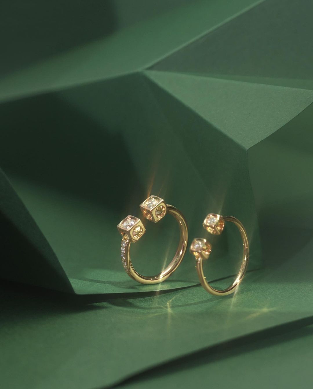 Close-up of two elegant gold rings with diamond cube detail on a dark green geometric surface. Jewelry product shot for fashion advertising.
