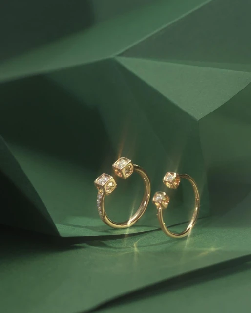 Close-up of two elegant gold rings with diamond cube detail on a dark green geometric surface. Jewelry product shot for fashion advertising.