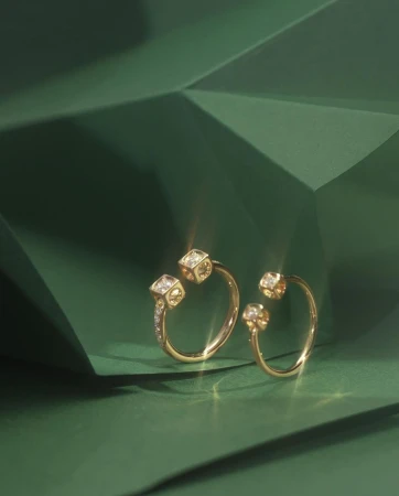 Close-up of two elegant gold rings with diamond cube detail on a dark green geometric surface. Jewelry product shot for fashion advertising.