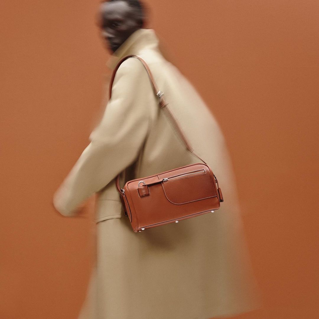Tan leather crossbody bag on a blurry figure in a tan coat against a tan background, product photography for a fashion advertisement.
