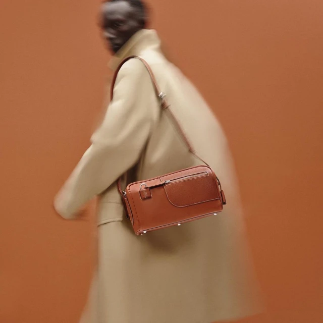 Tan leather crossbody bag on a blurry figure in a tan coat against a tan background, product photography for a fashion advertisement.