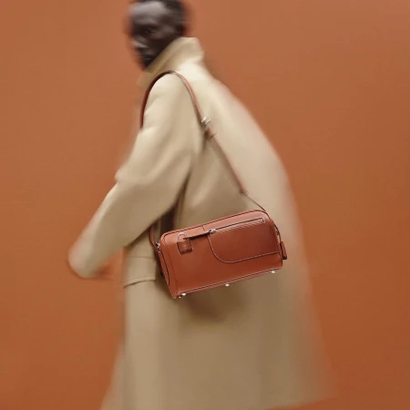 Tan leather crossbody bag on a blurry figure in a tan coat against a tan background, product photography for a fashion advertisement.