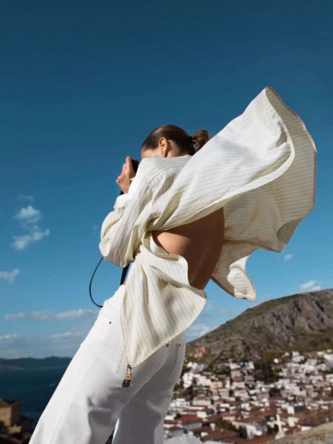 Young woman photographs a hillside village with a film camera. She wears a striped shirt, white pants, against a blue sky and mountain backdrop.