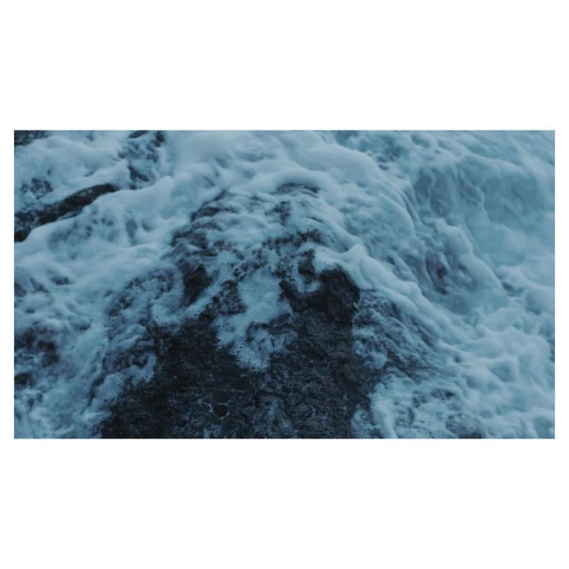 Close-up of ocean water crashing over dark rocks, creating a dynamic display of texture and motion in cool, muted tones.