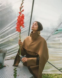 Model in a brown belted cape holding a tall red gladiolus flower inside a greenhouse