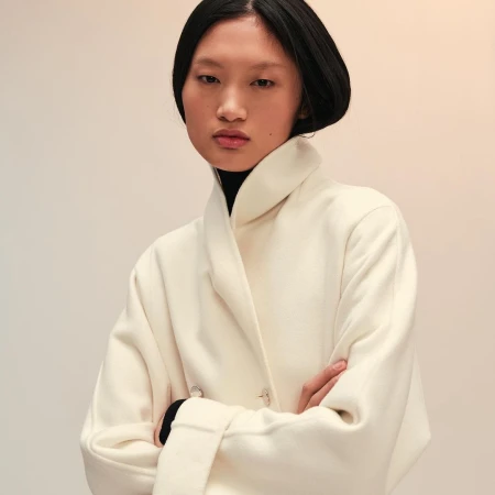East Asian woman with dark hair wearing a white coat with arms crossed against a plain gradient background
