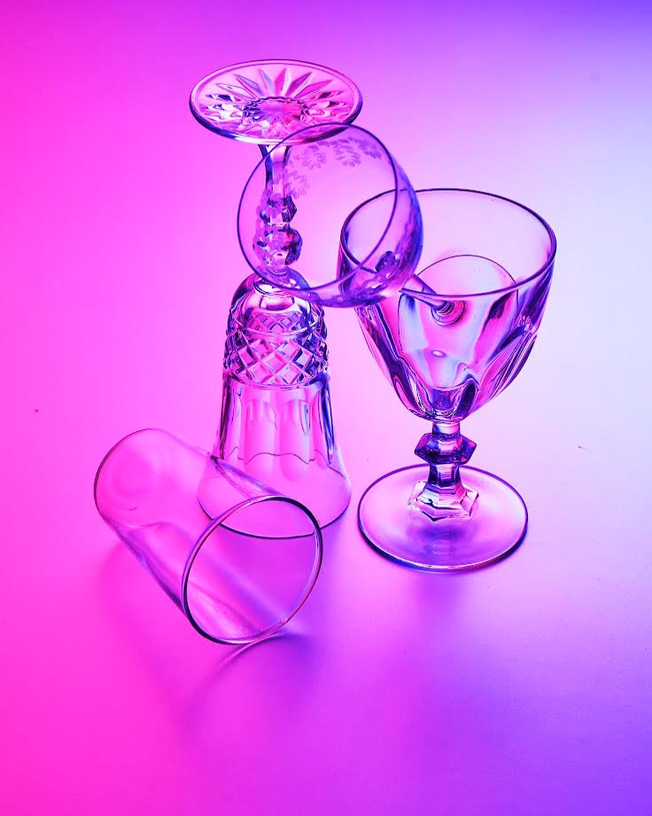 Studio shot of four clear crystal glasses stacked and lying down, illuminated with neon pink and blue lighting on a white background.