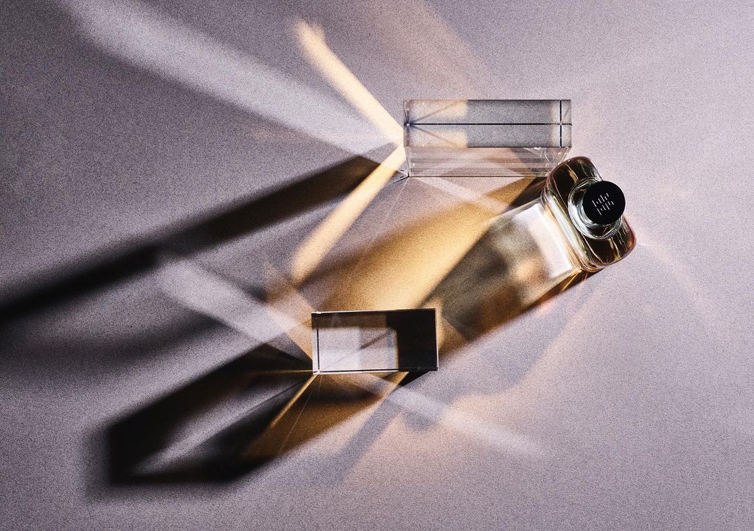 Top-down view of a clear glass perfume bottle with geometric transparent block on textured gray backdrop with sharp shadows and bright light.