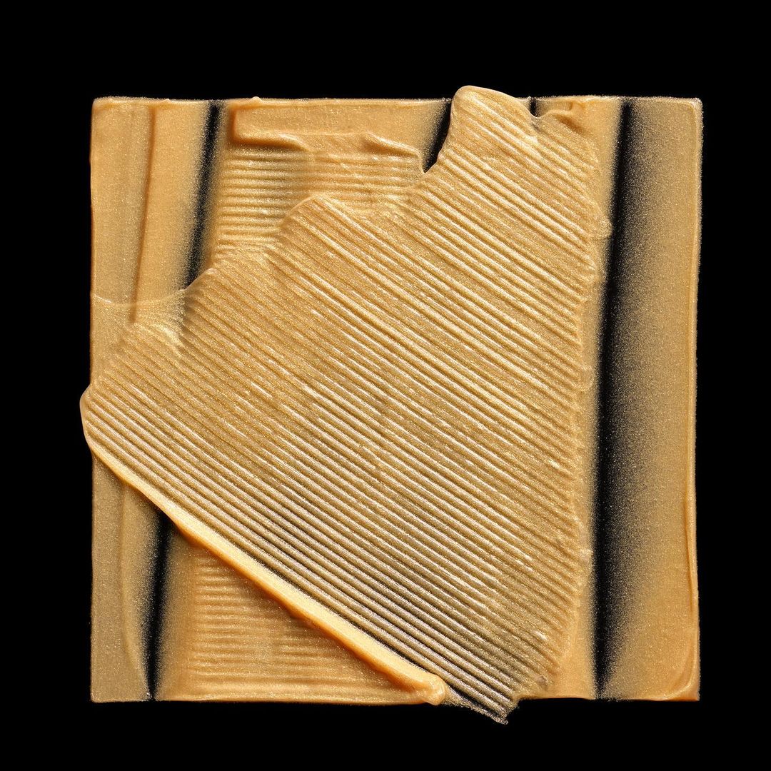 Close-up of a square bar of gold soap with grooved diagonal lines, set against a stark black background, highlighting texture.
