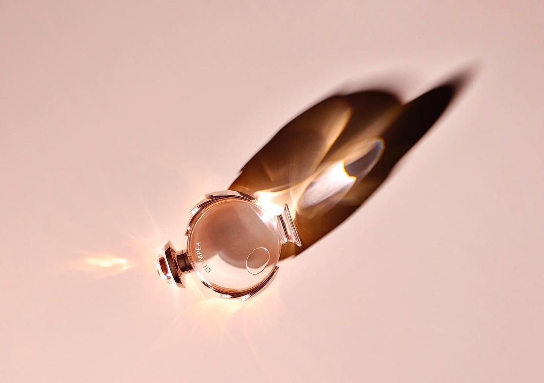 Overhead shot of a rose gold Olympea Paco Rabanne perfume bottle casting a diffused shadow on a light pink background.