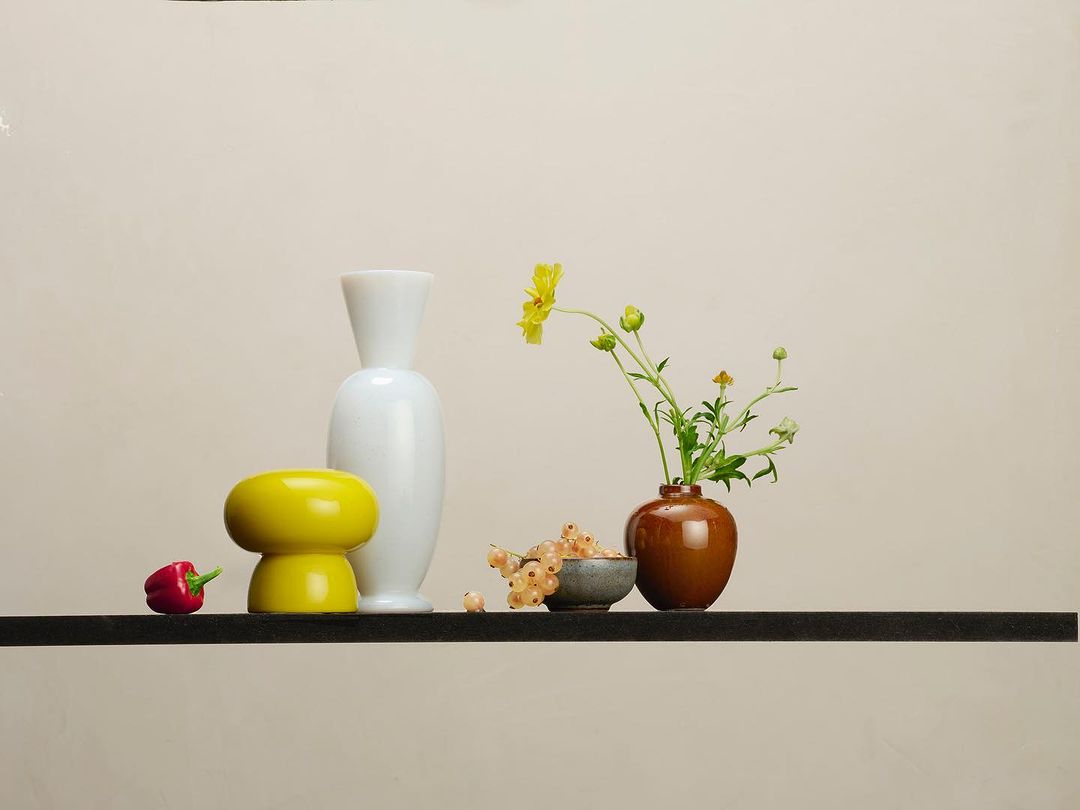 Still life with white and yellow vases, flowers with green stems in a brown vase, white currants, and a red pepper on a black shelf.