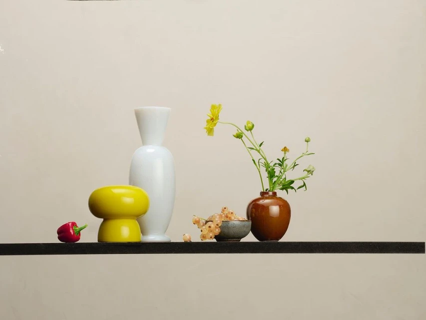 Still life with white and yellow vases, flowers with green stems in a brown vase, white currants, and a red pepper on a black shelf.