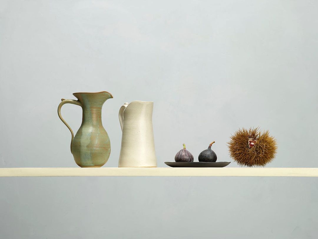 Still life featuring two ceramic pitchers, two figs on a small tray, and a chestnut bur, all arranged on a pale yellow surface.