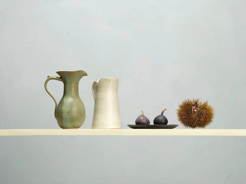 Still life featuring two ceramic pitchers, two figs on a small tray, and a chestnut bur, all arranged on a pale yellow surface.