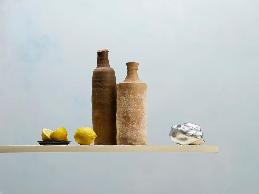 Still life featuring two clay bottles, lemons on a small plate, and a metallic rock on a light wooden shelf against a light blue backdrop.