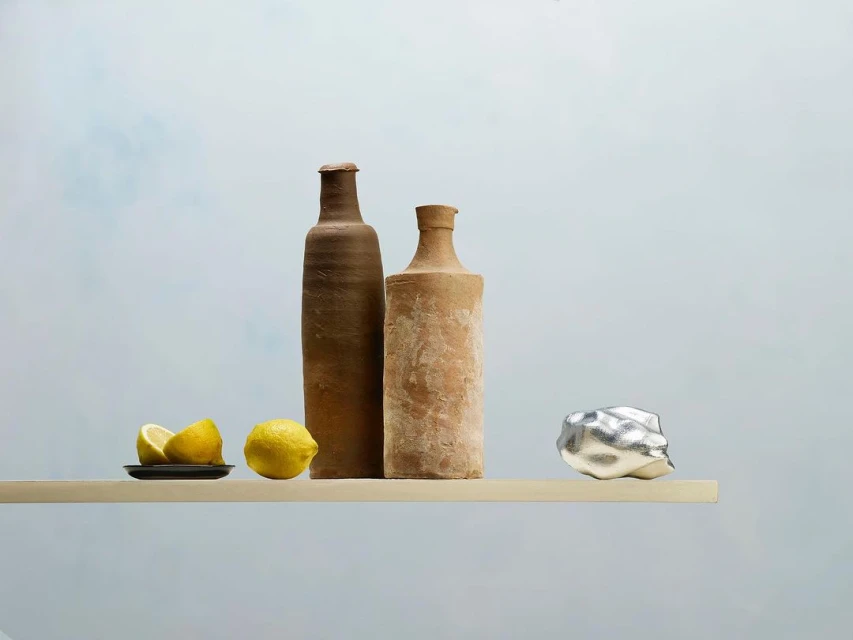 Still life featuring two clay bottles, lemons on a small plate, and a metallic rock on a light wooden shelf against a light blue backdrop.