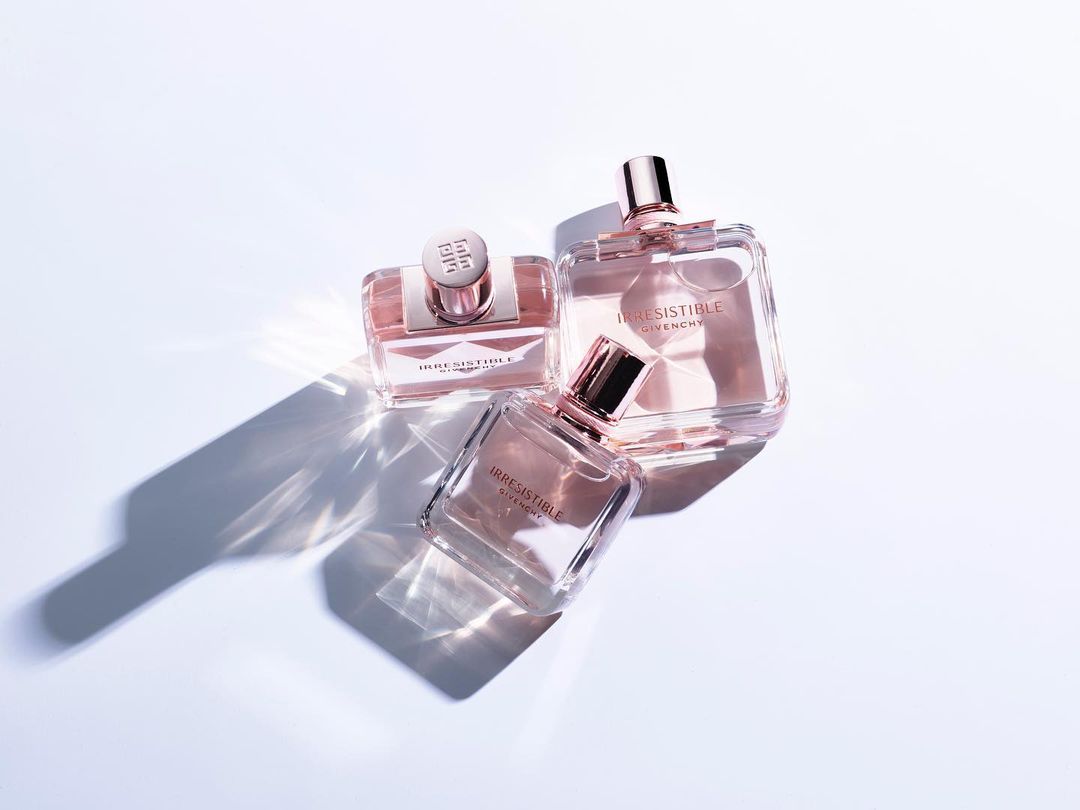 Three Givenchy Irresistible perfume bottles with rose gold accents are grouped on a white surface with light and shadow patterns.