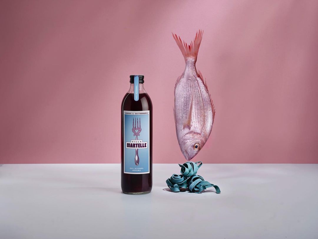 Still life with a bottle of red liquid, a raw fish, and teal pasta on a white surface against a light pink background.