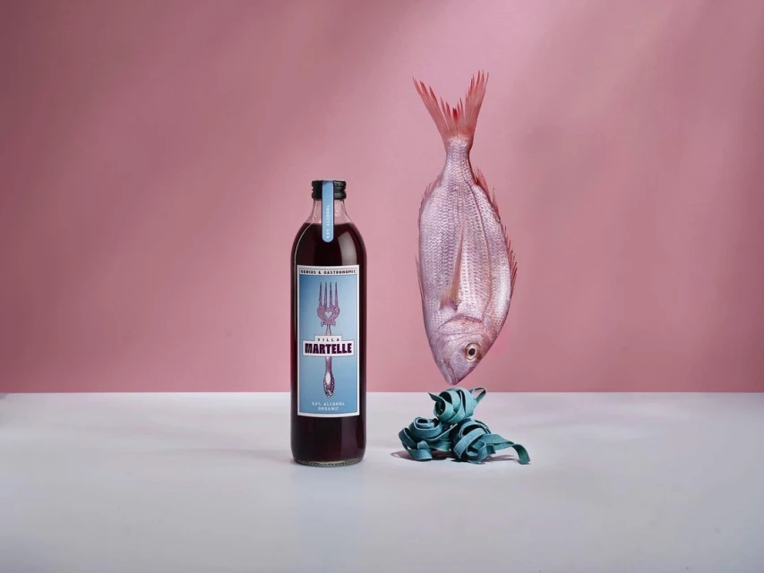 Still life with a bottle of red liquid, a raw fish, and teal pasta on a white surface against a light pink background.