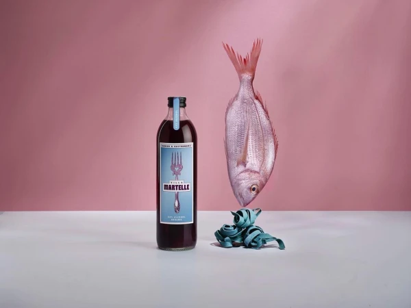 Still life with a bottle of red liquid, a raw fish, and teal pasta on a white surface against a light pink background.