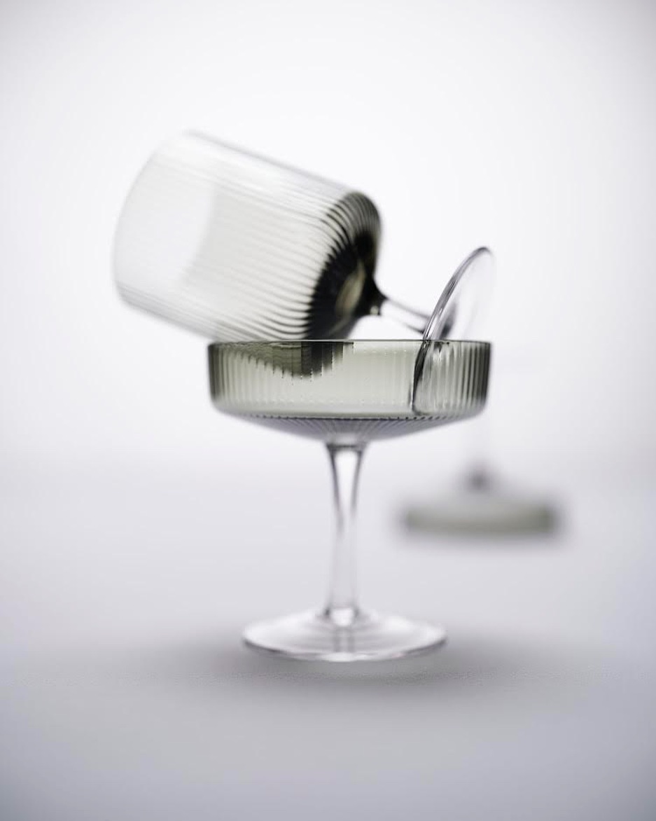 Two clear coupe glasses are stacked on a grey surface against a bright background. One glass is resting on the other.