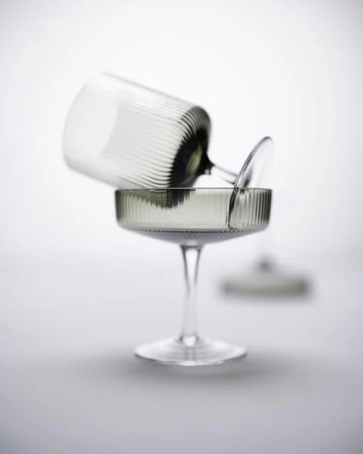 Two clear coupe glasses are stacked on a grey surface against a bright background. One glass is resting on the other.