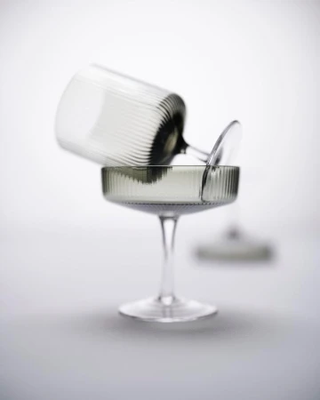 Two clear coupe glasses are stacked on a grey surface against a bright background. One glass is resting on the other.