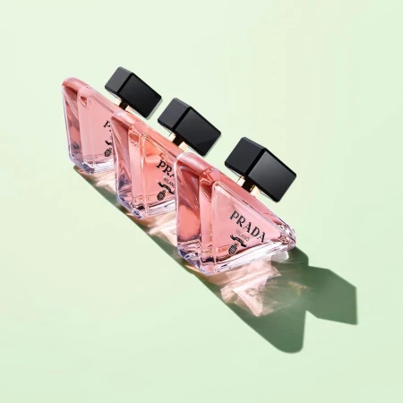 Studio shot of three Prada perfume bottles in a row, with triangular shape and black cap, casting shadows on a light green background.