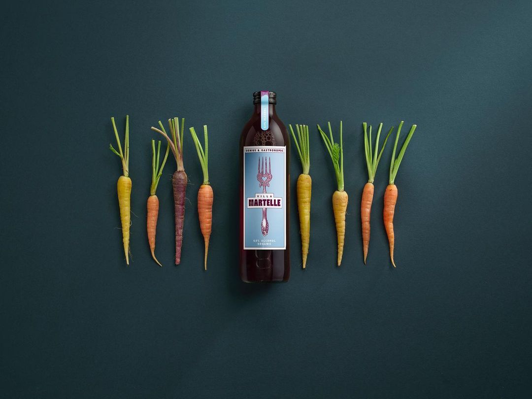Still life flat lay composition of raw carrots with green tops and a bottle of vegetable juice on a dark teal background.